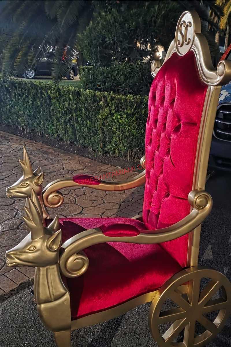 Red velvet Santa throne with gold reindeer armrests and carved wheels, set up outdoors in Miami
